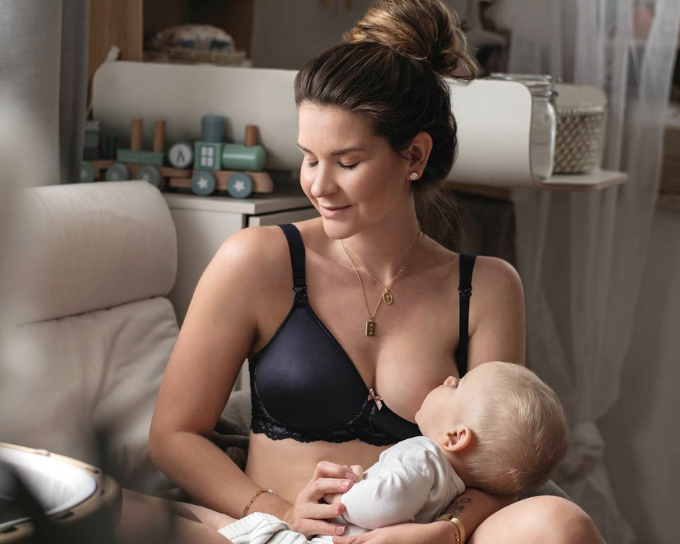 A sitting woman breastfeeding her baby while wearing the Anita maternity Miss Lovely wireless padded nursing bra in black